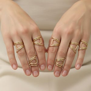 Op de afbeelding: Close-up van twee handen met meerdere goudkleurige ringen. De ringen hebben geometrische ontwerpen, waaronder driehoeken en gebogen lijnen. De vingers zijn kaal, behalve de ringen. De achtergrond is neutraal van kleur.