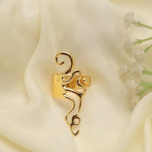 Könnte beinhalten: Ein goldfarbener Ring mit abstraktem Design. Der Ring hat ein breites Band und ein offenes Design mit geschwungenen und wirbelnden Formen. Der Ring liegt auf einem cremefarbenen Stoff.