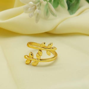 Puede incluir: Un anillo dorado con un diseño de doble banda. Cada banda presenta adornos en forma de hojas. El anillo se encuentra sobre un fondo amarillo suave, con un toque de flores blancas y hojas verdes en el fondo.