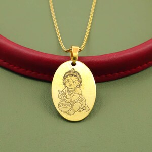 Puede incluir: Collar con colgante ovalado dorado con una imagen grabada de una figura sentada con corona, sosteniendo una flauta y una olla. El colgante cuelga de una fina cadena dorada. El fondo es rojo y verde.