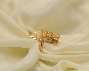 May include: A gold-colored ring featuring a detailed dragon head design. The dragon's head is intricately sculpted with open jaws and textured scales. The ring is set against a soft, cream-colored fabric, highlighting its ornate details.