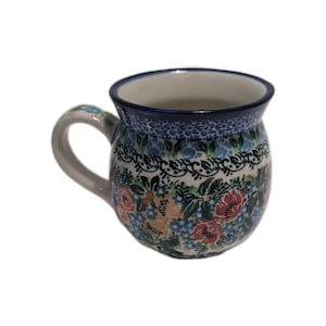Puede incluir: Una taza de cerámica con cuerpo redondeado y asa. La taza presenta un estampado floral con flores rojas, azules y amarillas sobre fondo blanco. El borde y el asa son azules.