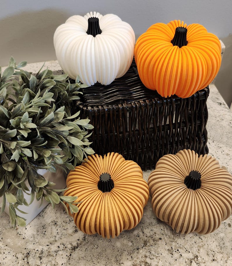 May include: A collection of decorative pumpkins in various colors, including white, orange, and tan. Each pumpkin has a ribbed design and a black stem. The pumpkins are arranged on a dark wicker basket and a speckled countertop.