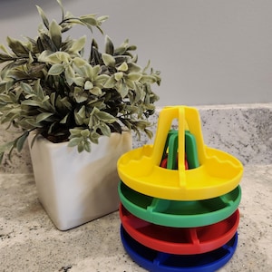 May include: A stackable toy with four colorful, circular trays in blue, red, green, and yellow. The toy has a central yellow support structure. A small, white square pot with a green plant is to the left.