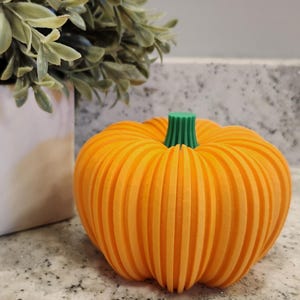 May include: An orange pumpkin with vertical grooves and a green stem. The decorative pumpkin is sitting on a speckled countertop next to a white pot with green leafy foliage. A fall-themed decoration.