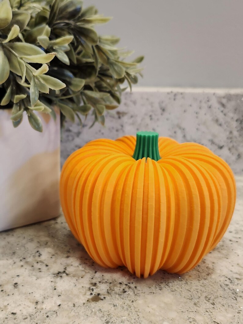 May include: An orange pumpkin with vertical grooves and a green stem. The decorative pumpkin is sitting on a speckled countertop next to a white pot with green leafy foliage. A fall-themed decoration.