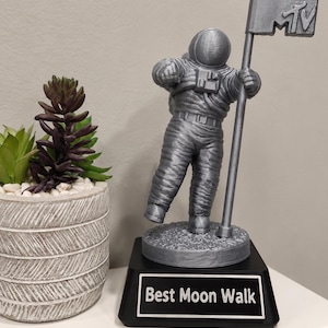 May include: A silver-colored astronaut figurine standing on a circular base, holding a pole with an MTV flag. The figure is mounted on a black rectangular trophy base with a plaque that reads "Best Moon Walk" in white text.