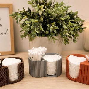 May include: Three oval-shaped bathroom organizers in dark brown, gray, and terracotta orange. Each container features two compartments, one holding white cotton swabs and the other holding a stack of round white cotton pads for facial care.