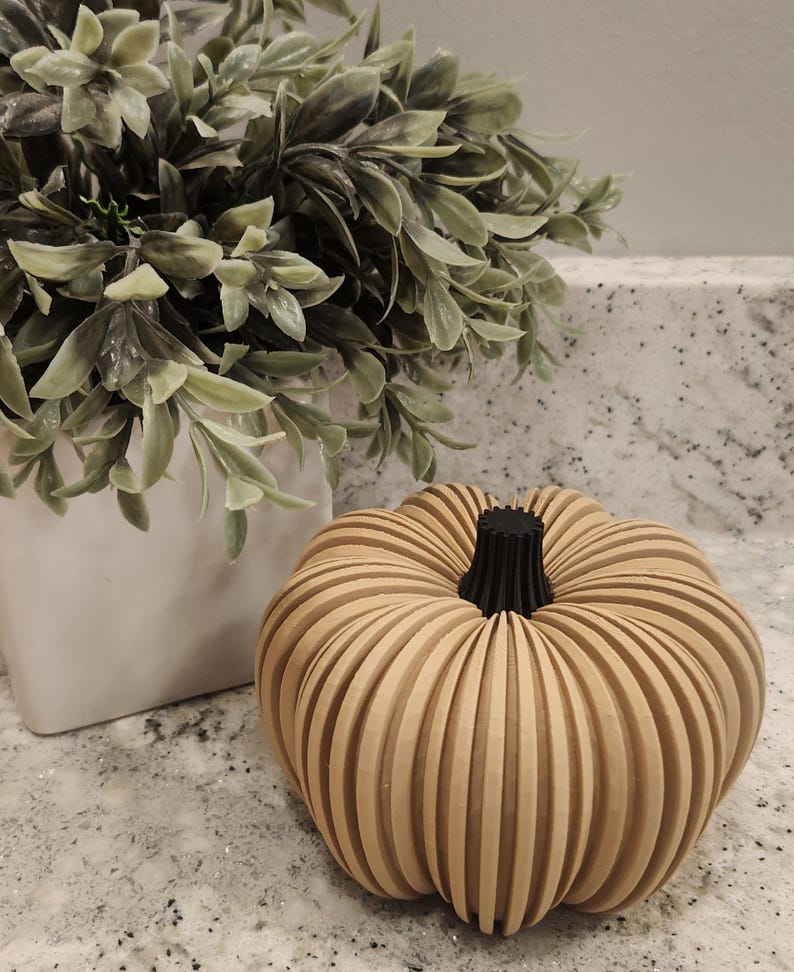 May include: A decorative wooden pumpkin with a black stem. The pumpkin is light brown and has vertical grooves. A white vase with green leafy plants is in the background. The pumpkin is a fall-themed decoration.