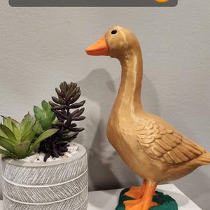 May include: A golden goose figurine with an orange beak and feet, standing on a green base. Above the goose, a dark gray rectangular label displays the white text Golden Goose followed by a smiling face emoji with heart-shaped eyes.