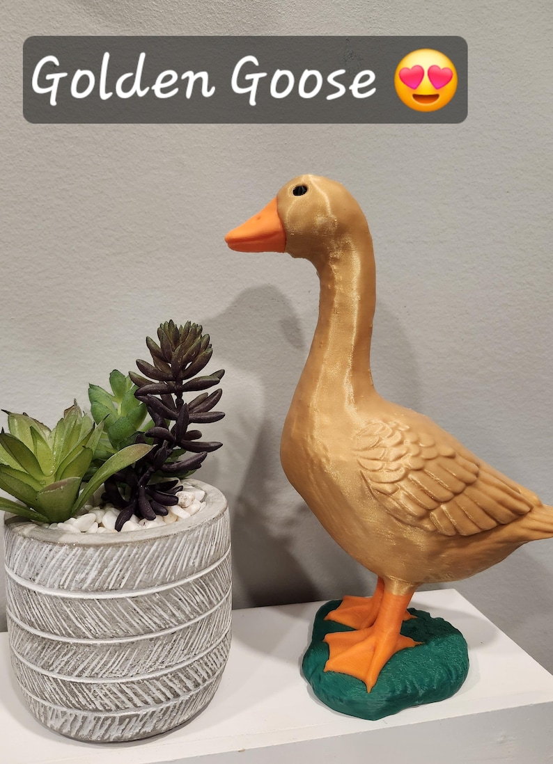May include: A golden goose figurine with an orange beak and feet, standing on a green base. Above the goose, a dark gray rectangular label displays the white text Golden Goose followed by a smiling face emoji with heart-shaped eyes.