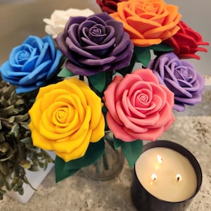 May include: A bouquet of artificial roses in vibrant colors including yellow, pink, purple, blue, orange, red, and white, featuring green leaves on stems. A three-wick candle in a dark container sits nearby, emitting a warm glow from its flames.