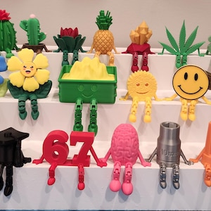 May include: A collection of small, 3D-printed figurines with articulated legs, featuring various designs including a skull, a graduation cap, the number 67, a brain, a 10mm socket, a wire nut, a pineapple, a smiley face, and cacti.
