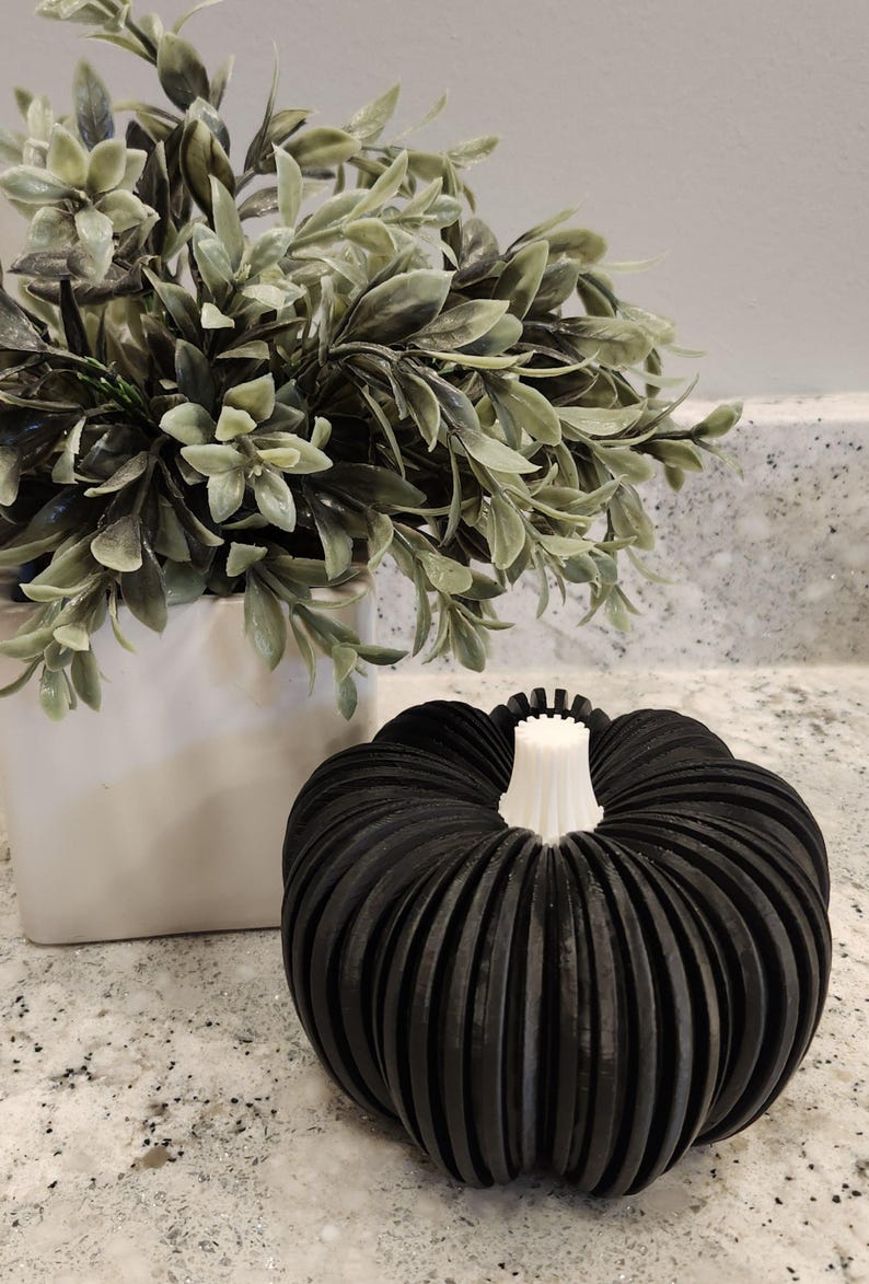 May include: A black, ribbed pumpkin decoration with a white stem sits on a countertop. A white square pot with green leafy plants is in the background. The pumpkin is a fall-themed decorative item.
