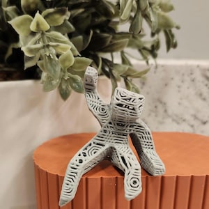 May include: A gray, 3D-printed figurine of a person in a dynamic pose, with arms raised. The figure is placed on a small, round, orange-brown pedestal. A green plant is in the background.