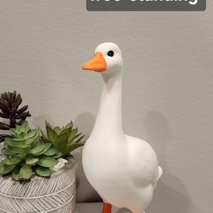 May include: A white goose figurine with an orange beak and orange webbed feet. The goose is free-standing and measures 7 inches in height as indicated by the text overlay. The figure has a smooth matte finish and black eyes.