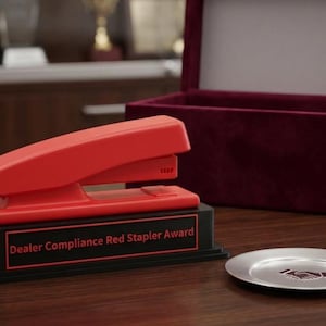May include: A bright red stapler mounted on a black rectangular base with a plaque that reads Dealer Compliance Red Stapler Award. Next to the base is a small silver plate featuring a handshake icon, and behind it sits an open maroon velvet box.