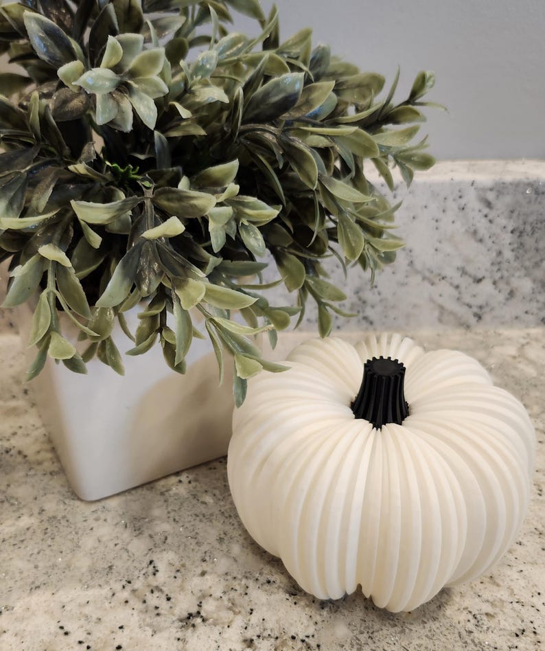 May include: A white, ribbed pumpkin with a black stem sits on a speckled surface. A square white planter with green foliage is in the background. The pumpkin is a decorative item, perfect for fall.