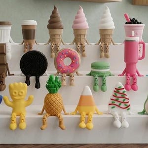 May include: A collection of small, articulated figurines shaped like various food and drink items, including ice cream cones, a chocolate bar, a donut, a pineapple, a candy corn, and an avocado, all arranged on a white tiered display stand.