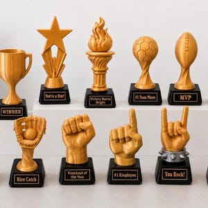 May include: A collection of twelve gold-colored trophies with black bases, featuring various designs including a cup, star, flame, sports balls, and hand gestures. Each trophy has a small plaque with text such as Winner, MVP, and You Rock.