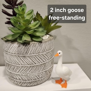 May include: A white goose figurine with orange feet and beak, standing next to a gray cylindrical planter with a textured herringbone pattern containing green and dark purple artificial succulents. The text 2 inch goose free-standing is visible.