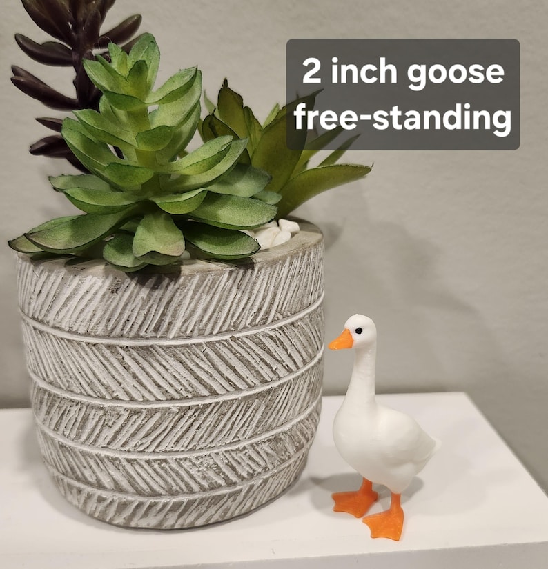 May include: A white goose figurine with orange feet and beak, standing next to a gray cylindrical planter with a textured herringbone pattern containing green and dark purple artificial succulents. The text 2 inch goose free-standing is visible.