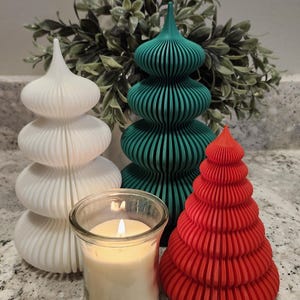 May include: A selection of decorative Christmas tree-shaped objects in white, teal, and red. A lit candle in a glass jar is in the foreground. The trees have a ribbed design. Green foliage is in the background.