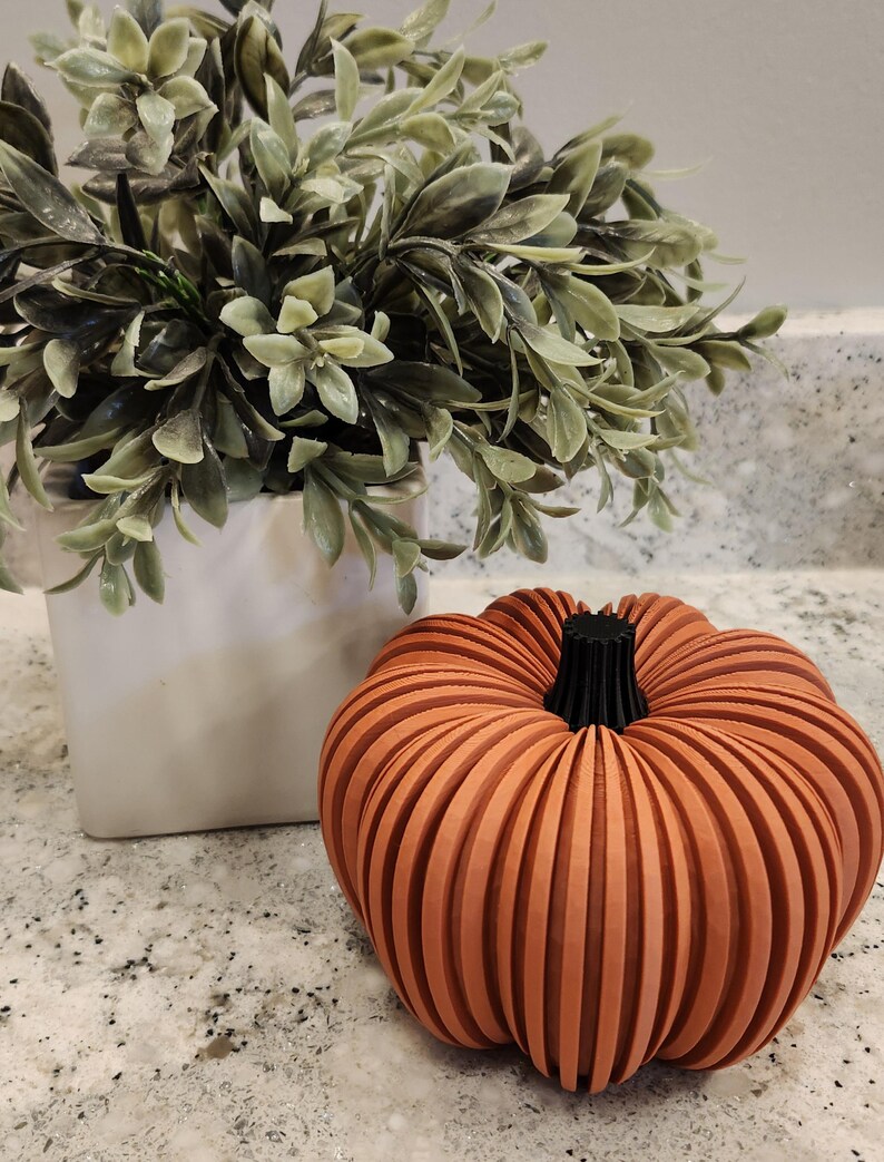 May include: A decorative orange pumpkin with vertical grooves and a black stem. Beside it is a white square vase with green leafy plant. The pumpkin is a fall-themed decoration.