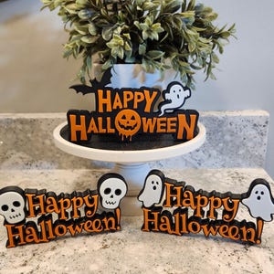 May include: Halloween decorations featuring the phrase "Happy Halloween!" in black and orange. The designs include a bat, a ghost, a jack-o'-lantern, and skulls. One decoration is on a white cake stand, and two are on a countertop.