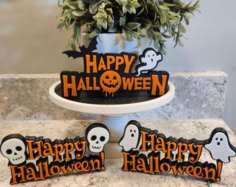 May include: Halloween-themed decorations with the words "Happy Halloween!" in black and orange. The decorations include a bat, a ghost, a jack-o'-lantern, and skulls. One decoration is on a white cake stand, and two are on a countertop.