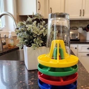 May include: A stack of four circular plastic drying racks in yellow, green, red, and blue, each featuring a central support structure. A clear glass tumbler is inverted and placed over the top yellow rack to facilitate drying.