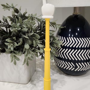 May include: A yellow foam finger with a white hand pointing upwards. The foam finger is on a white surface, with a plant in a white pot and a navy blue decorative vase in the background.