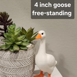 May include: A white goose figurine with an orange beak and orange webbed feet. The goose is free-standing and measures 4 inches in height. The text 4 inch goose free-standing is displayed in a dark gray box above the goose.
