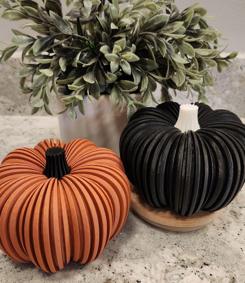 May include: Two decorative pumpkins, one orange and one black, sit on a countertop. The orange pumpkin has a black stem, and the black pumpkin has a white stem. Both pumpkins have a ribbed design. A green plant in a silver pot is in the background.