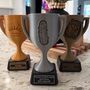 May include: Three plastic trophies with black bases. The gold trophy features a chef hat and reads 'SNACK ATTACK 2026 1st Place Snack of All Snacks'. The silver trophy features a pickle and reads 'SNACK ATTACK 2026 2nd Place Still a Big Dill'.