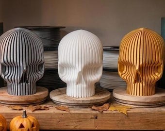 May include: Three skull sculptures arranged in a row, each featuring a vertical ribbed design. The left skull is metallic gray, the center is matte white, and the right is metallic gold. Each skull rests on a circular wooden base.