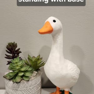 May include: A white goose figurine with an orange beak and orange feet, standing on a green base. The text 7 inch goose standing with base is visible at the top. The goose has black eyes and is positioned next to a gray textured planter.