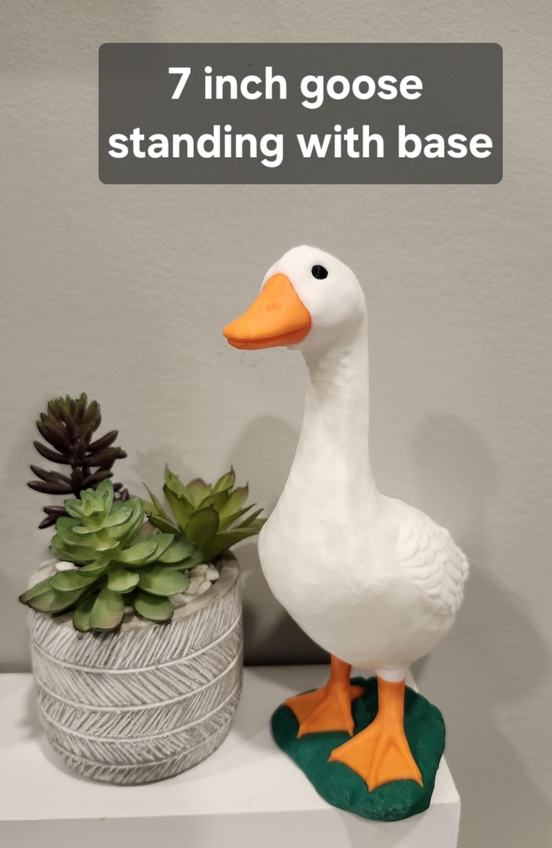 May include: A white goose figurine with an orange beak and orange feet, standing on a green base. The text 7 inch goose standing with base is visible at the top. The goose has black eyes and is positioned next to a gray textured planter.