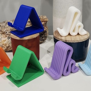 May include: A selection of colourful phone stands in various shapes and designs. The stands are in shades of orange, green, blue, purple, and white. The stands are made of plastic and have a textured surface.