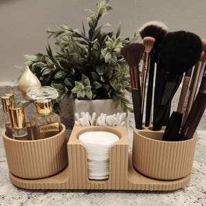 Peut inclure: Un organisateur de cosmétiques beige et côtelé avec trois sections. Une section contient des flacons de parfum, une autre des cotons, et la troisième des pinceaux de maquillage. Une petite plante est en arrière-plan.