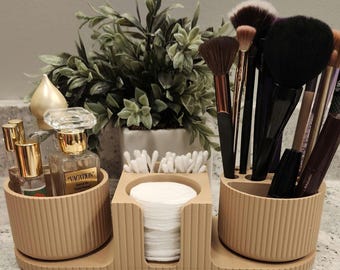 Puede incluir: Un organizador de cosméticos beige y acanalado con tres secciones. Una sección contiene frascos de perfume, otra discos de algodón y la tercera brochas de maquillaje. Una pequeña planta está en el fondo.