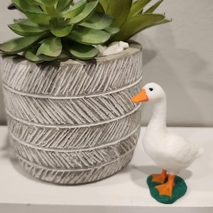 May include: A white goose figurine with an orange beak and feet, standing on a green base, labeled 2 inch goose standing with base. Next to it is a gray cylindrical planter with a textured herringbone pattern containing green succulents.