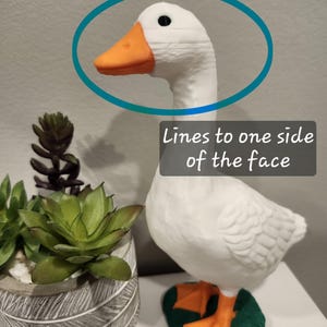 May include: A white goose figurine with an orange beak and orange webbed feet standing on a green base. The goose has a black eye and visible horizontal printing lines on one side of its face. The text "Lines to one side of the face" is present.