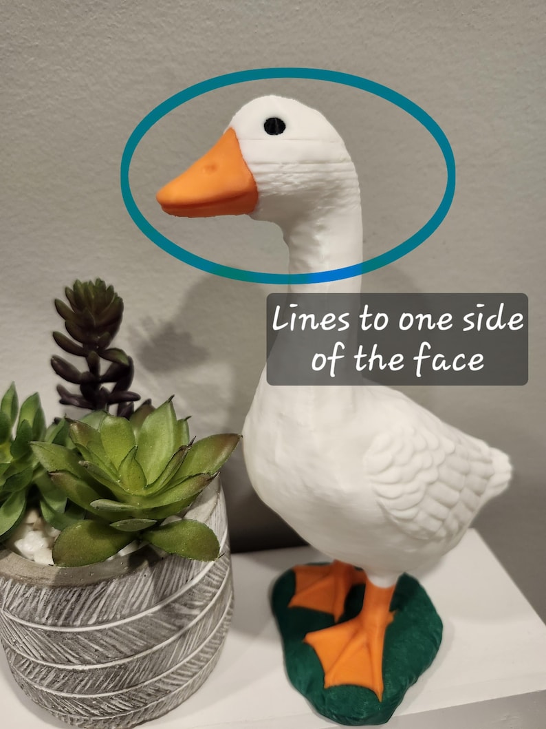 May include: A white goose figurine with an orange beak and orange webbed feet standing on a green base. The goose has a black eye and visible horizontal printing lines on one side of its face. The text "Lines to one side of the face" is present.
