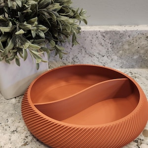 May include: A terracotta-colored, round bowl with a divider, featuring a textured, diagonal ribbed design on the outer edge. The bowl sits on a speckled countertop, with a white pot holding greenery in the background.