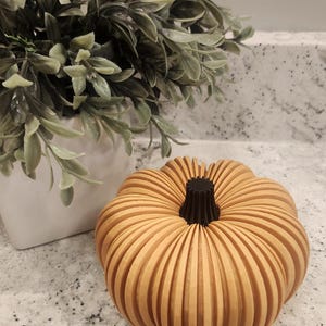 May include: A wooden pumpkin with a light orange color and a dark brown stem. The pumpkin is made of vertical wooden slats, giving it a ribbed appearance. A white pot with green leaves is in the background.
