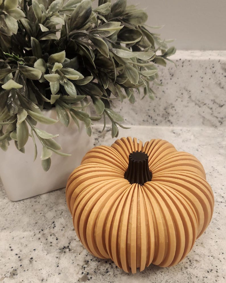 May include: A wooden pumpkin with a light orange color and a dark brown stem. The pumpkin is made of vertical wooden slats, giving it a ribbed appearance. A white pot with green leaves is in the background.