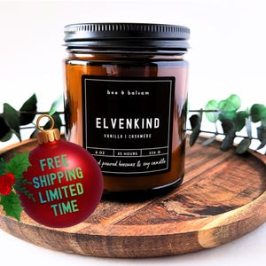 May include: A brown glass jar candle with a black lid, labeled "ELVENKIND" with "Vanilla & Cashmere" scent. The candle is on a wooden tray, decorated with greenery and a red Christmas ornament.