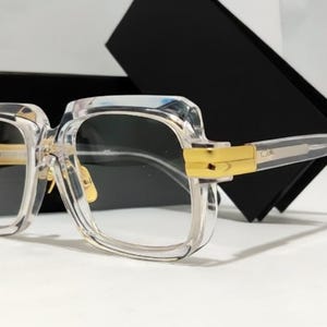 May include: Clear square-framed eyeglasses featuring thick transparent acetate rims and polished gold-tone metal hinges at the temples. The glasses have a prominent bridge and clear arms, presented with a black rectangular storage case behind them.