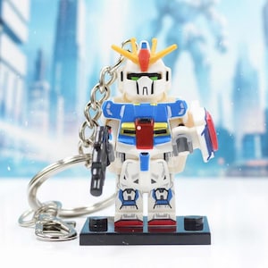 May include: A Gundam robot keychain featuring a white, blue, and red plastic figure with a yellow V-fin crest on its head. The figure holds a black handheld weapon and is attached to a silver metal chain and ring, standing on a black base.